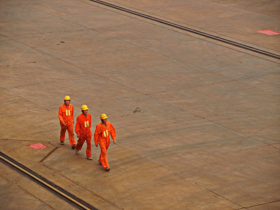 Three workers in hard hats and orange coveralls walking across a paved yard